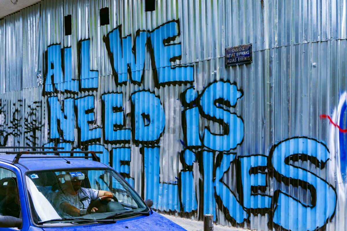 Graffiti on a corrugated metal wall reads ALL WE NEED IS LIKES in large blue and black letters—a playful nod to the impact of a strong video marketing strategy. A blue car with people inside appears in the foreground, below a small street sign.