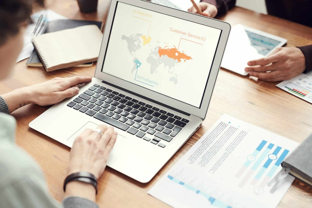 A person engaged in web based training uses a laptop displaying a world map with business metrics, seated at a wooden table filled with notebooks, papers, and another person’s hands nearby.