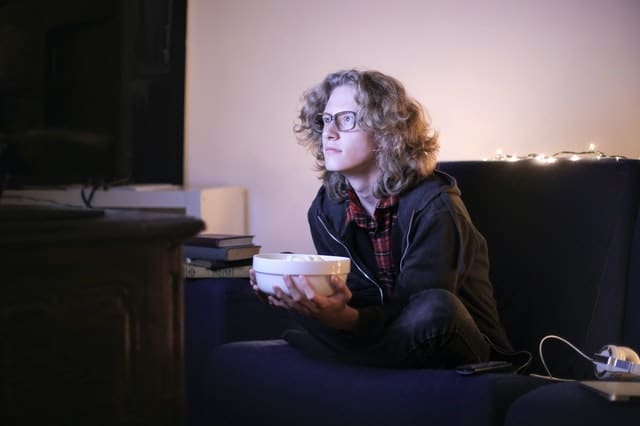 blonde teen sits on the couch with a bowl of popcorn in his hands and watches a streaming ad