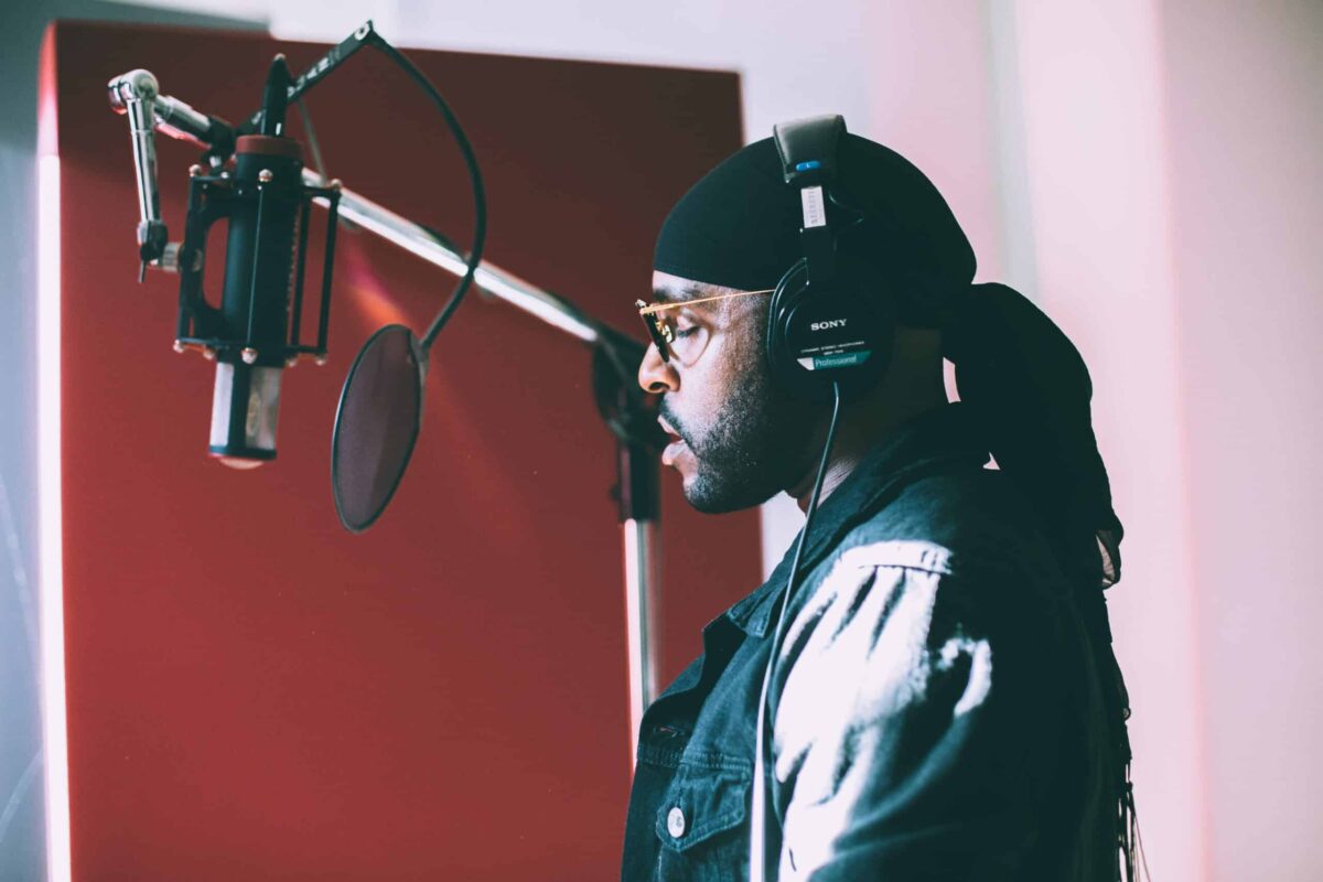A man wearing headphones and a black headscarf stands in front of a microphone in a recording studio, appearing focused as he records vocals for music licensing for video projects.