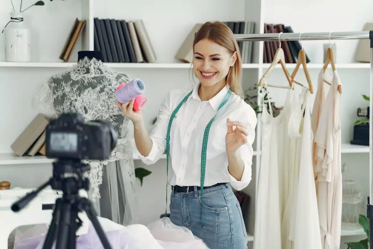 A smiling woman with a measuring tape around her neck holds spools of thread, confidently being on camera in a room filled with dresses, hangers, and bookshelves—likely filming a fashion or sewing tutorial.