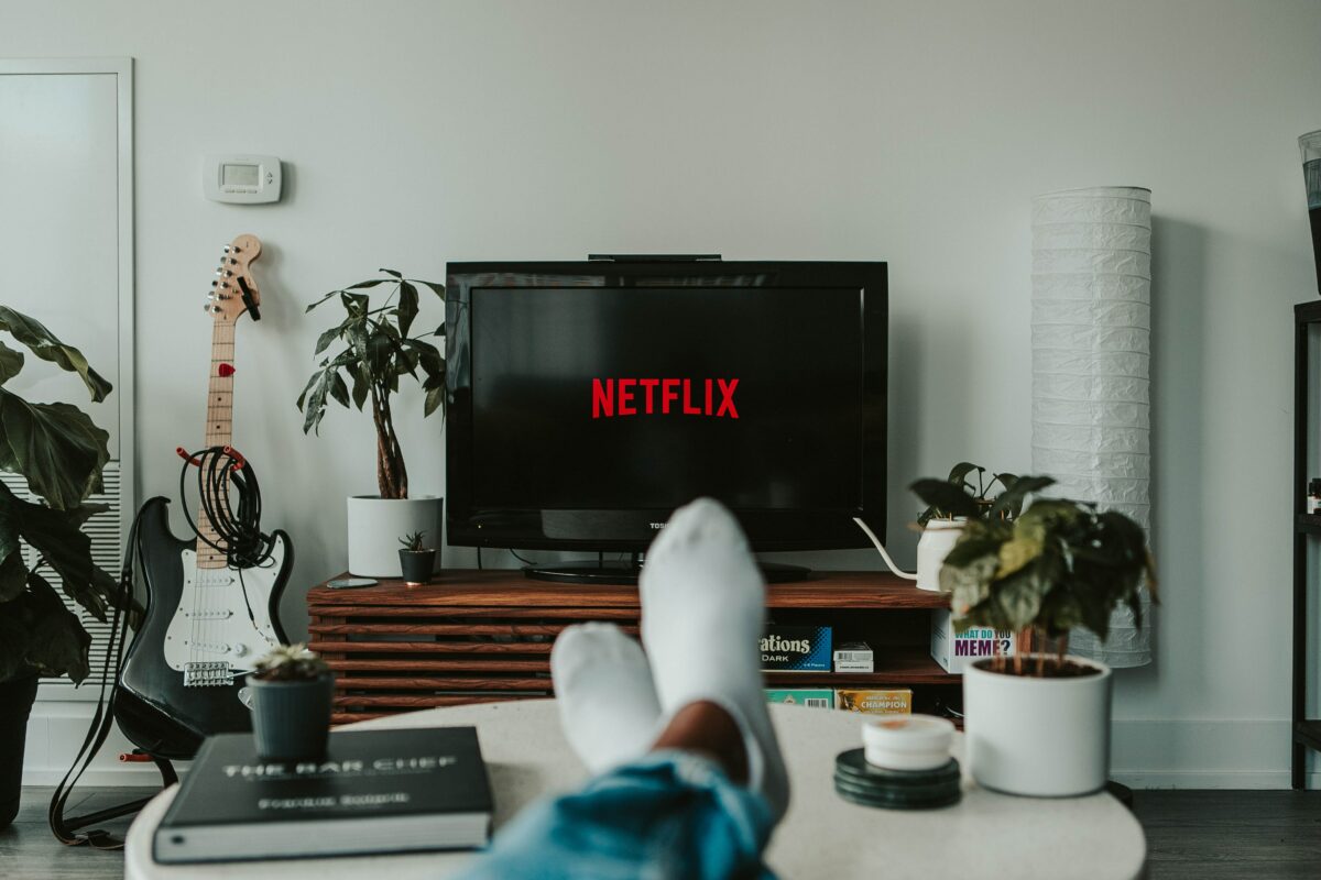 A person with their feet up on a table faces a TV displaying the Netflix logo in a cozy living room, pondering where to advertise amidst plants, a guitar, and books scattered around the inviting space.