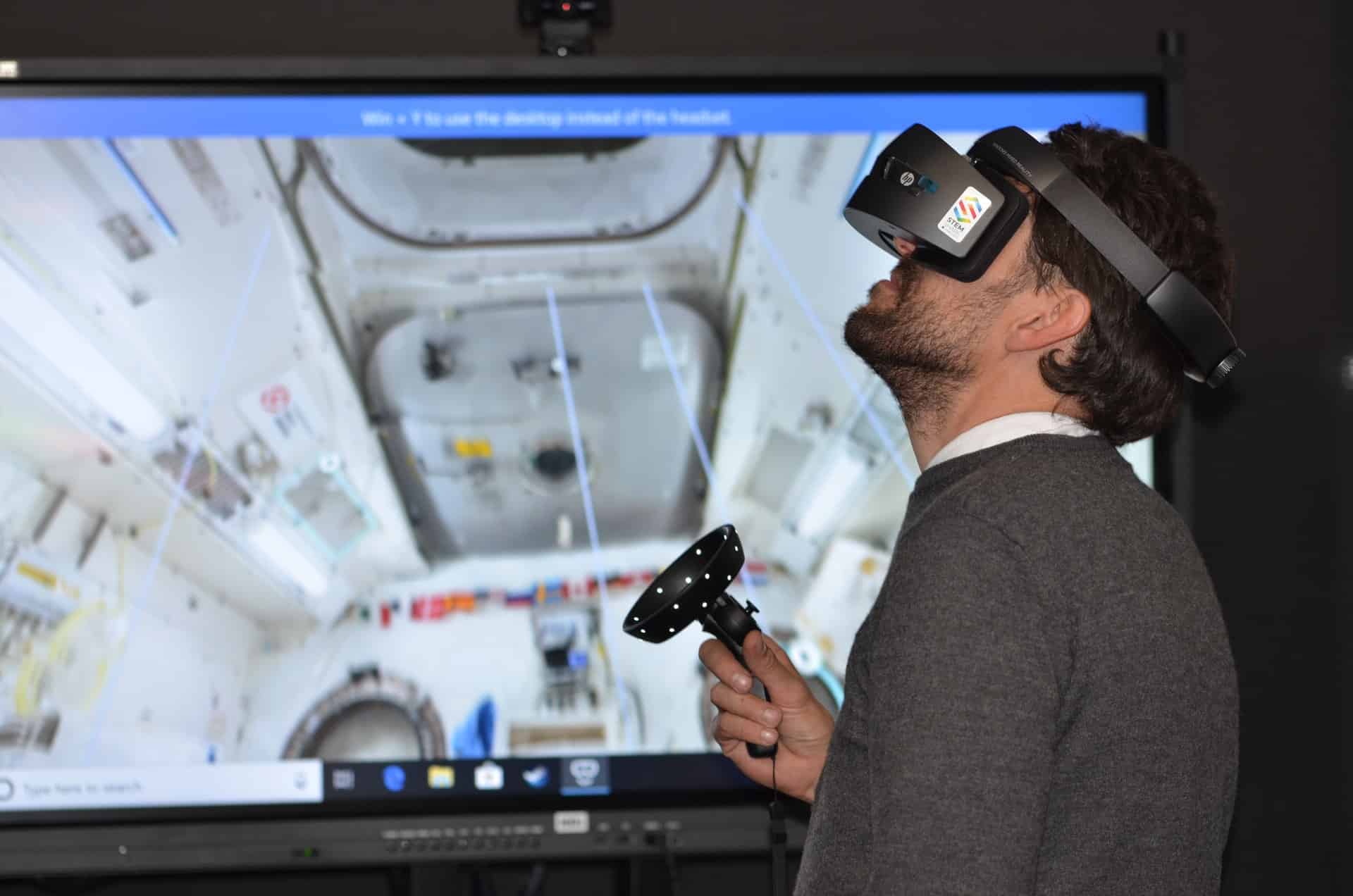 White man with a beard wears a VR headset and holds a controller while standing in front of a screen showing a space capsule.