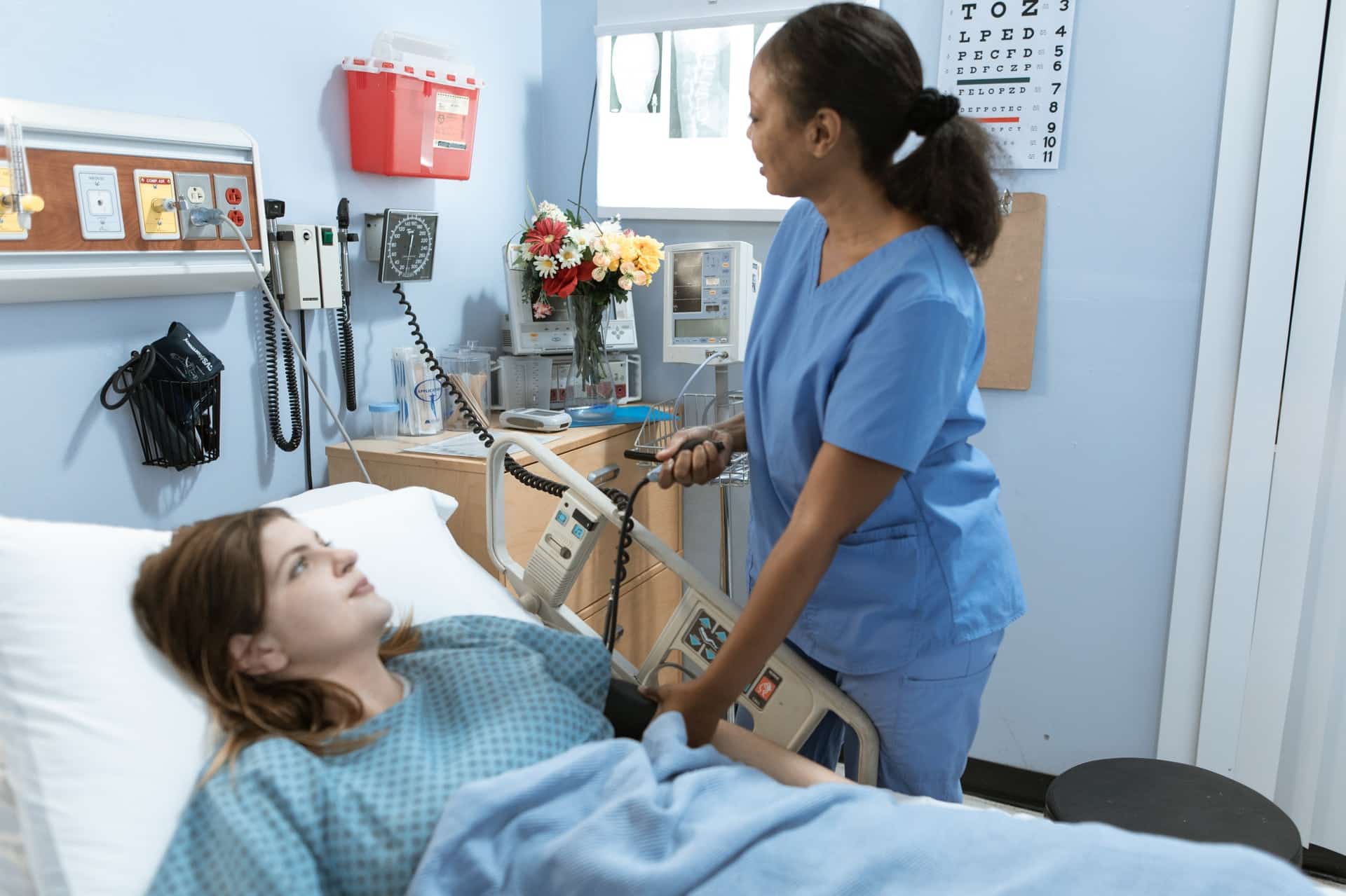 a white female patient in a hospital bed has her blood pressure checked by a black nurse wearing blue scrubs