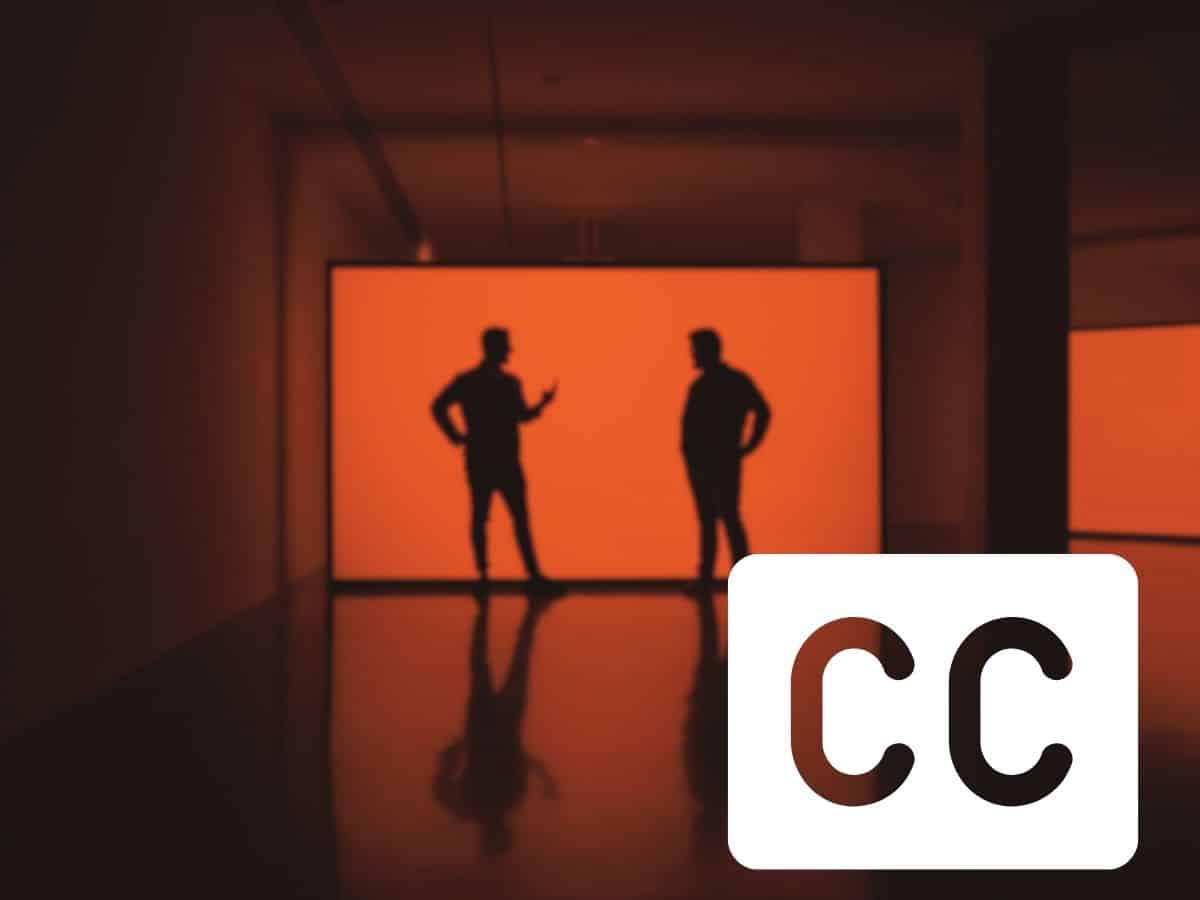 Image of two men standing against an orange background. A white closed captions symbol appears in the bottom right corner