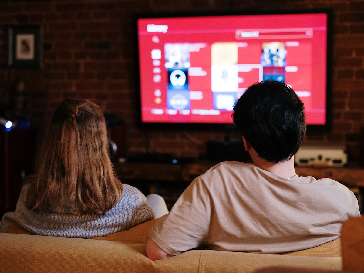 a couple sit on a couch with their backs to the camera. They are watching OTT video on their connected TV