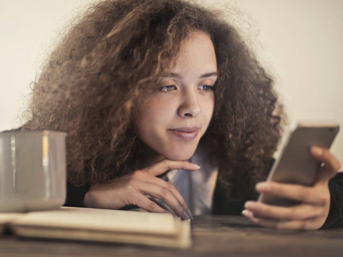 A black woman leans forward toward a desk while looking at her phone. Her hand is on the desk in front of her.
